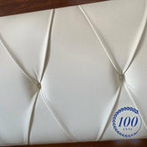 Dettaglio della testata imbottita di un letto matrimoniale in vera pelle colore bianco, con raffinata lavorazione capitonné a bottoni. Il bordo superiore e inferiore della testata è incorniciato in legno di ciliegio, in perfetta armonia con lo stile classico della collezione. In basso a destra è visibile il logo del centenario Piombini (1925–2025) con la scritta “100 anni” circondata da una corona d’alloro.